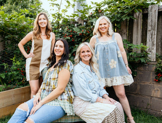 Four women posing together outdoors with greenery and a wooden bench in the background.